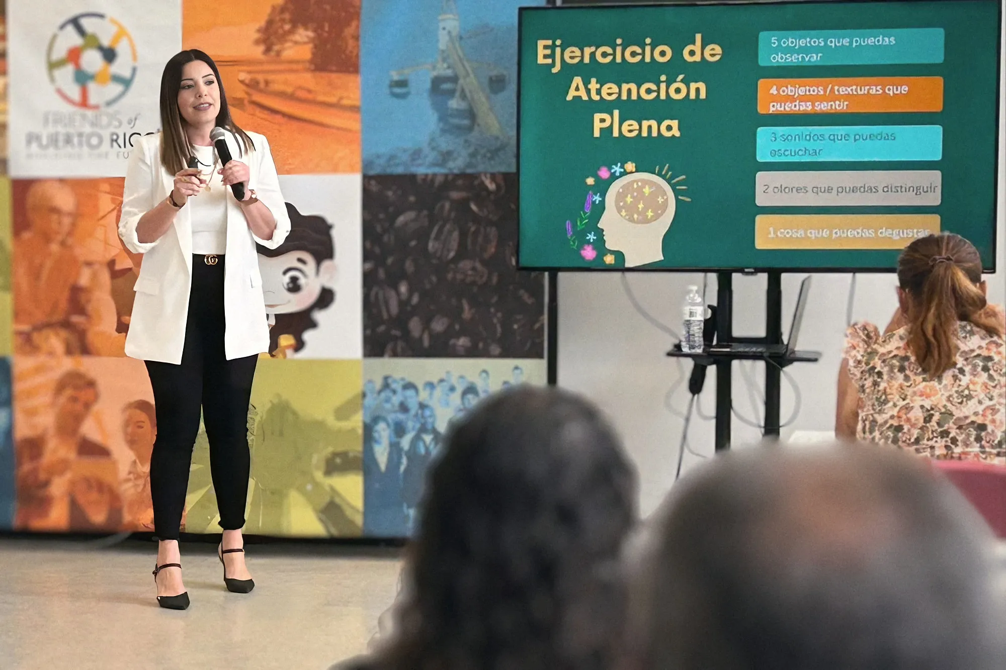 Melanie, de pie con micrófono frente a un grupo, guía un ejercicio de atención plena proyectado en pantalla